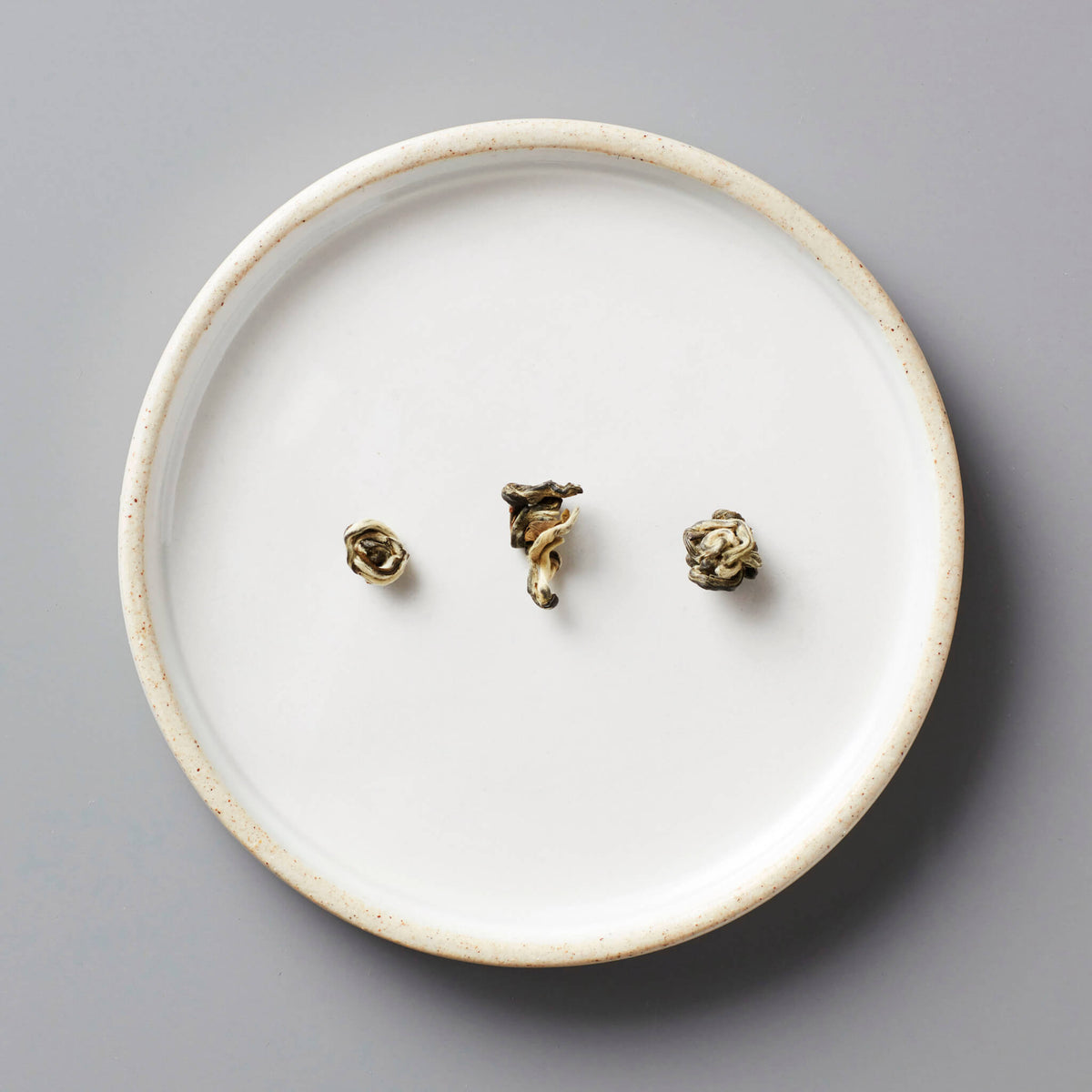 Round white ceramic plate with speckled rim holding three tightly rolled tea pearls of Jasmine Bai Hao, a scented green tea from Guangxi, China.