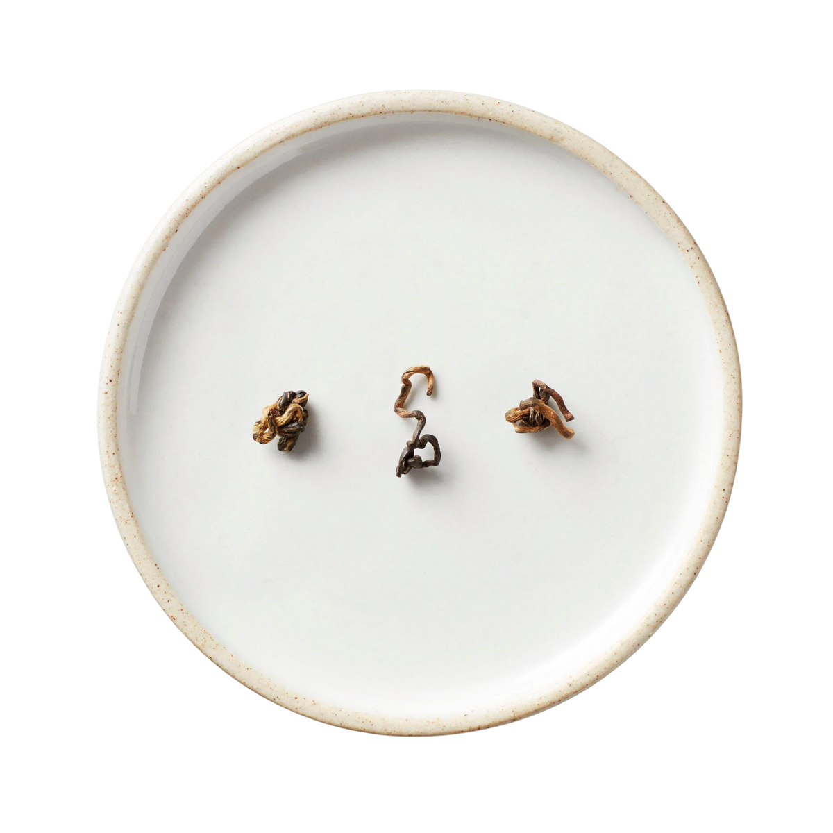 White ceramic plate displaying twisted, brown-and-gold hand-rolled tea leaves, showcasing Hugo Tea's Gao Wen black tea from Yunnan, distinguished by its high-heat roasting process.