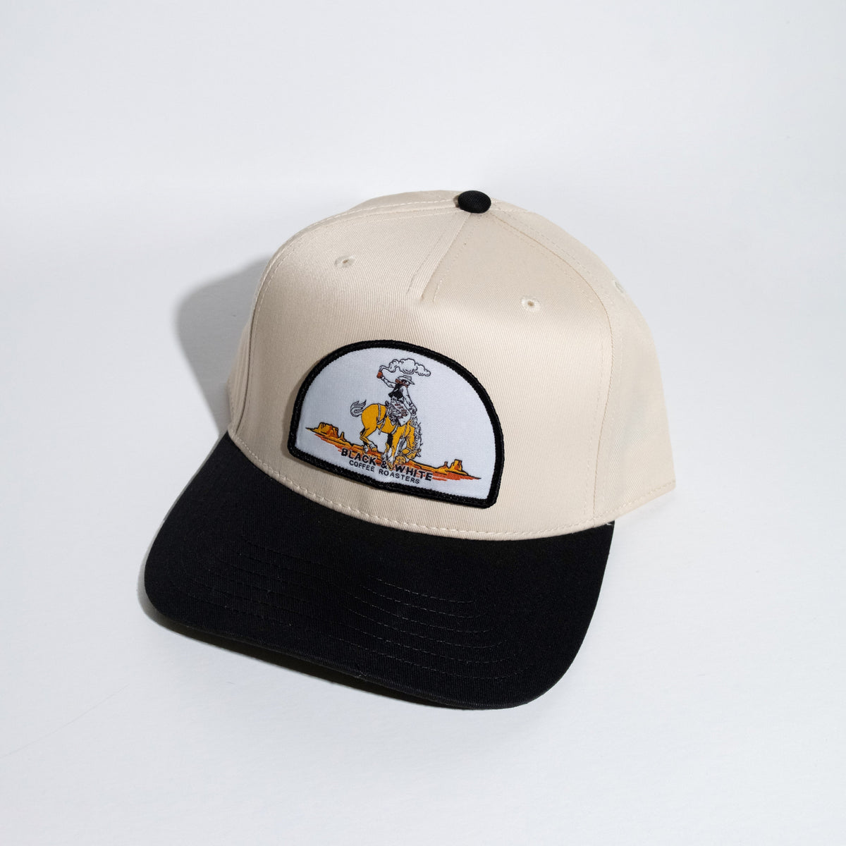 A structured baseball cap with a cream-colored crown and a black flat brim, featuring a circular embroidered patch on the front with a cowboy cartoon character.