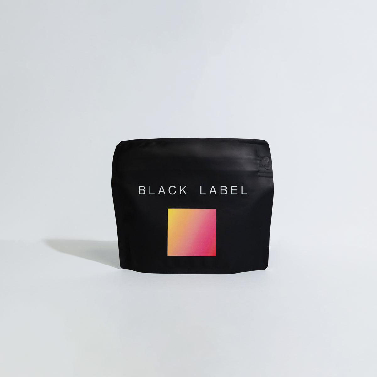 A black coffee bag with the text 'BLACK LABEL' and a small square gradient label in yellow, orange, and pink, set against a plain light gray background.
