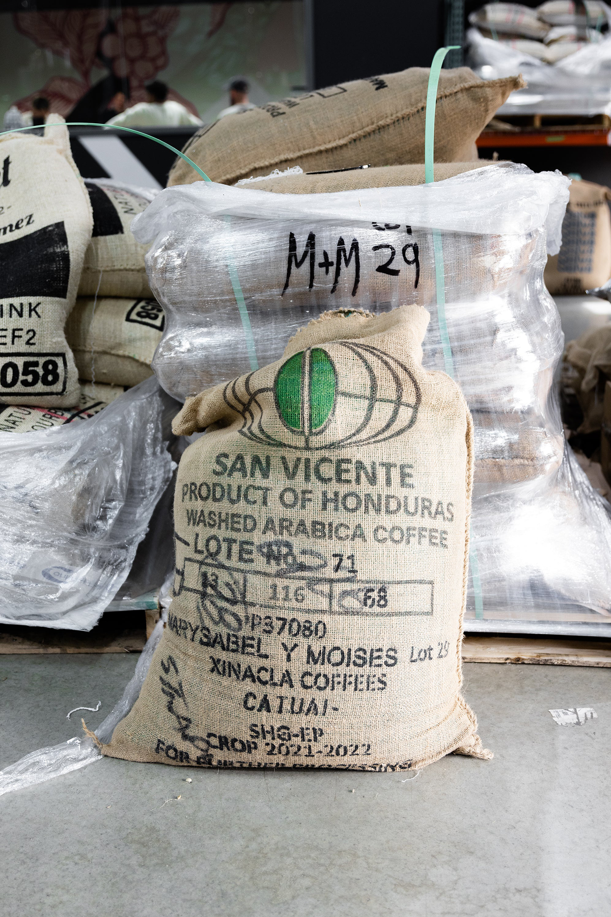 A bag of green coffee in a jute bag sitting on the floor of a warehouse with the words, San Vincente, Product of Honduras washed Arabica coffee on them.