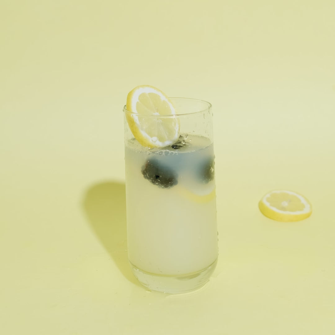 A video of a glass of lemonade with lemons and blackberries floating in it and a hand coming into frame and setting a slice of lemon on the edge of the glass. 