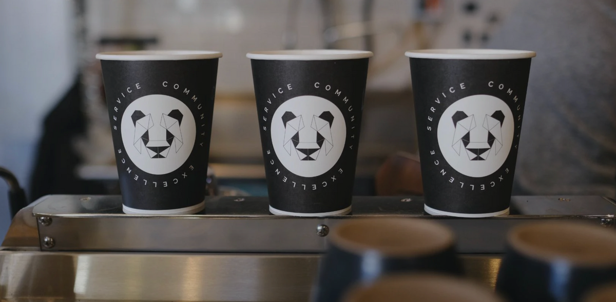A stack of paper coffee cups with a geometric panda in a white circle with black triangle eyes and black ears. The whole design is symmetrical  and made of various triangles. Around the white circle are the words Service, community and excellence. 